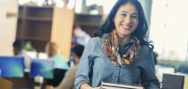 Mid adult Hispanic woman is standing at checkout counter in college library. She's holding stack of library text books. Adult student is wearing casual clothing and scarf. Other students are sitting at computers in background. Library is modern with bright windows and tall bookshelves.