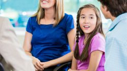 Pretty young girl in a pink shirt with brown hair sits at a meeting with her mom and brother.