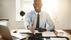 Smiling young African executive extending his arm in a handshake while sitting at his desk in an office