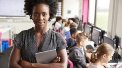 Portrait Of Female Teacher Holding Digital Tablet Teaching Line Of High School Students Sitting By Screens In Computer Class