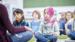 A diverse group of preschool children are sitting on the ground cross-legged while meditating. They are practicing mindfulness as they face their teacher.