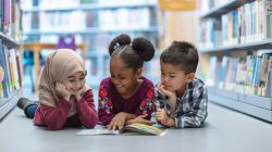 Three (3) kids who are friends are laying on the floor in between bookshelves in the library. They are reading a book together. They are all smiling and enjoying their afternoon.